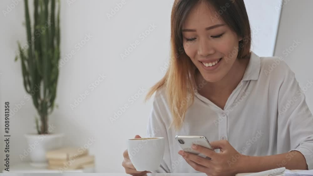 Smiling asian business woman using her smartphone while holding a cup of tea or coffee in the white office Smiling asian business woman using her smartphone while holding a cup of tea or coffee in the white office
