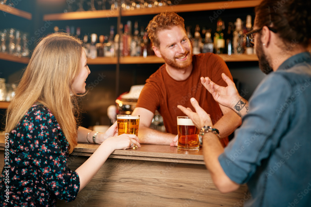 Cheerful friends leaning on bar counter, drinking beer and chatting with bartender. Night out.