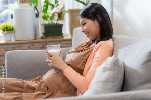 Asian pregnant woman hold a glass of milk for her health and baby with smile and sitting on the sofa, feeling happily and relaxed. Pregnant women concept.