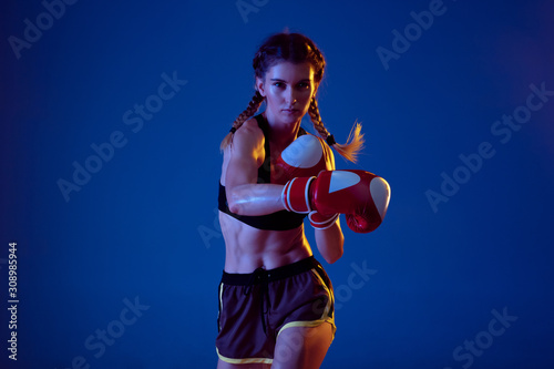 On the run. Fit caucasian woman in sportswear boxing on blue studio background in neon light. Novice female caucasian boxer working out and training. Sport, healthy lifestyle, movement concept.