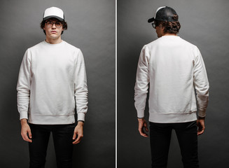 Man wearing blank white sweatshirt and empty baseball cap standing over gray background. Sweatshirt or hoodie for mock up, logo designs or design print with with free space.