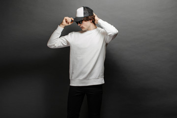 Man wearing blank white sweatshirt and empty baseball cap standing over gray background. Sweatshirt or hoodie for mock up, logo designs or design print with with free space.