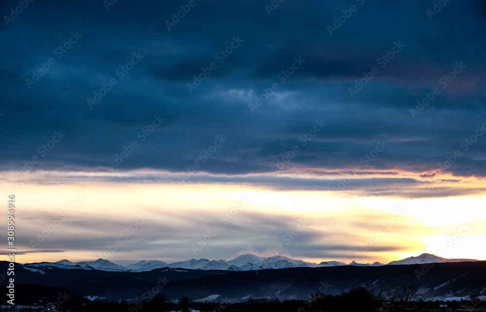 Sunset over the snow-capped mountain peaks of the Caucasus in cloudy weather in winter. The rays of the sun at sunset break through the clouds.