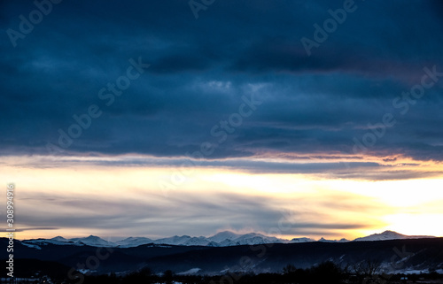 Fototapeta Naklejka Na Ścianę i Meble -  Sunset over the snow-capped mountain peaks of the Caucasus in cloudy weather in winter. The rays of the sun at sunset break through the clouds.