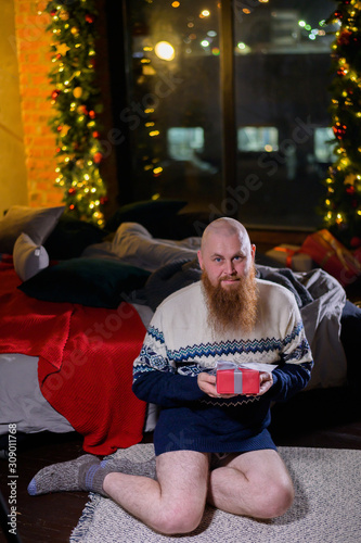 A man with a long red beard sits on the floor without pants in a winter sweater. man is holding a wrapped present on the background of New Year decorations and lights. Christmas tree. Parody, humor.