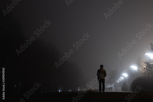 Fototapeta Naklejka Na Ścianę i Meble -  A man standing with his back to us in a jacket on an evening alley in the fog illuminated by evening white lamps against a background of birches and shrubs.