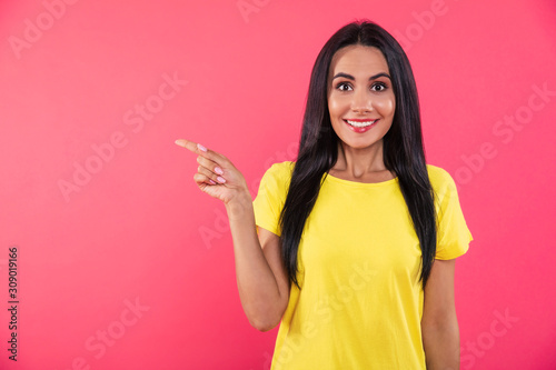 You bet! Close-up photo of a stunning happy girl, who is pointing to the left with her right index finger and posing in a yellow t-shirt on a pink background.