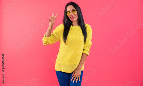 Flourishing youth. Young happy woman in a yellow knitted sweater and sunglasses is posing in front, showing a peace sign with her right hand and smiling.
