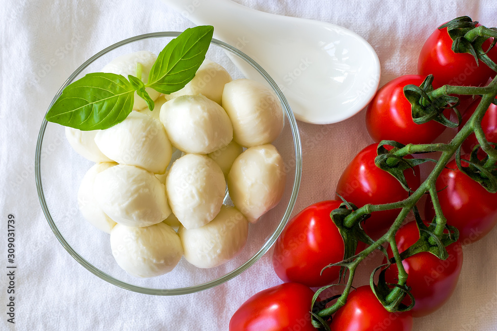 Mini mozzarella cheese balls in glass bowl and branch of red cherry tomatoes on a white linen background. Healhy mediterranean cuisine and vegetarian food.