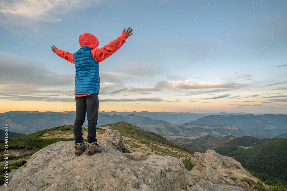 Young child boy hiker standing with raised hands in mountains enjoying view of amazing mountain landscape at sunset.