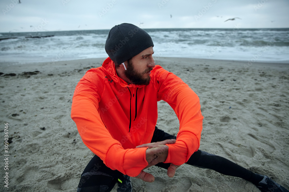 Photo of young dark haired pensive bearded sportsman dressed in warm athletic clothes doing stretching exercises, preparing for morning workout over coastline of sea