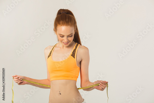 beautiful young woman with red hair in sportswear measures her waist on a white background © Alexandr