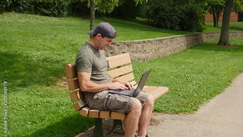 Middle aged young man sitting on a bench in the park recieves bad news and is stressed while working on his laptop computer.
