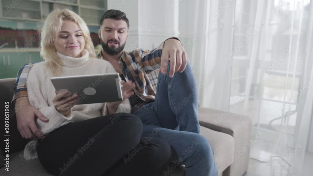 Portrait of young Caucasian family choosing purchase with the use of digital tablet. Handsome man and beautiful woman doing online shopping at home. E-commerce, technologies, wireless, internet. Portrait of young Caucasian family choosing purchase with the use of digital tablet. Handsome man and beautiful woman doing online shopping at home. E-commerce, technologies, wireless, internet.