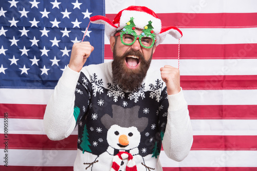 Christmas traditions and observances changed greatly over time. American bearded hipster guy joined cheerful celebration. American tradition. Santa Claus on american flag. Winter holidays season © be free