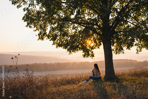 Attractive young girl in casual clothes sitting under big tree with book and looking at the beautiful sunset