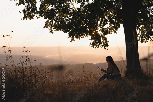 Young female student sitting under big tree and reading a book, beautiful landscape on the backgriud