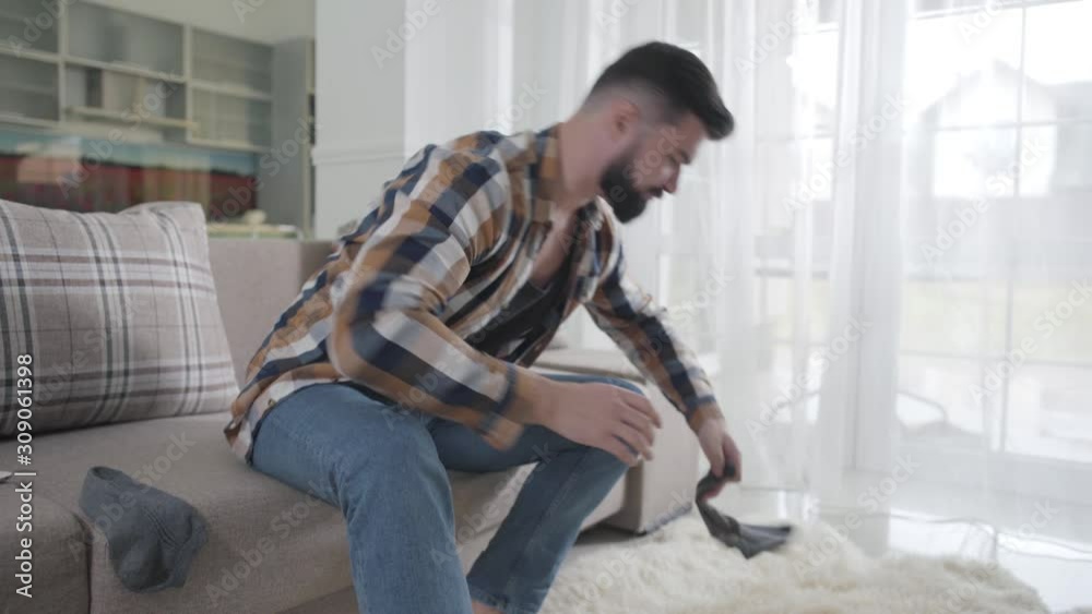 Bearded Caucasian man looking for clean socks as sitting on couch. Guy sniffing sock he found under the sofa, putting it on and leaving. Getting dressed, single man. Bearded Caucasian man looking for clean socks as sitting on couch. Guy sniffing sock he found under the sofa, putting it on and leaving. Getting dressed, single man.