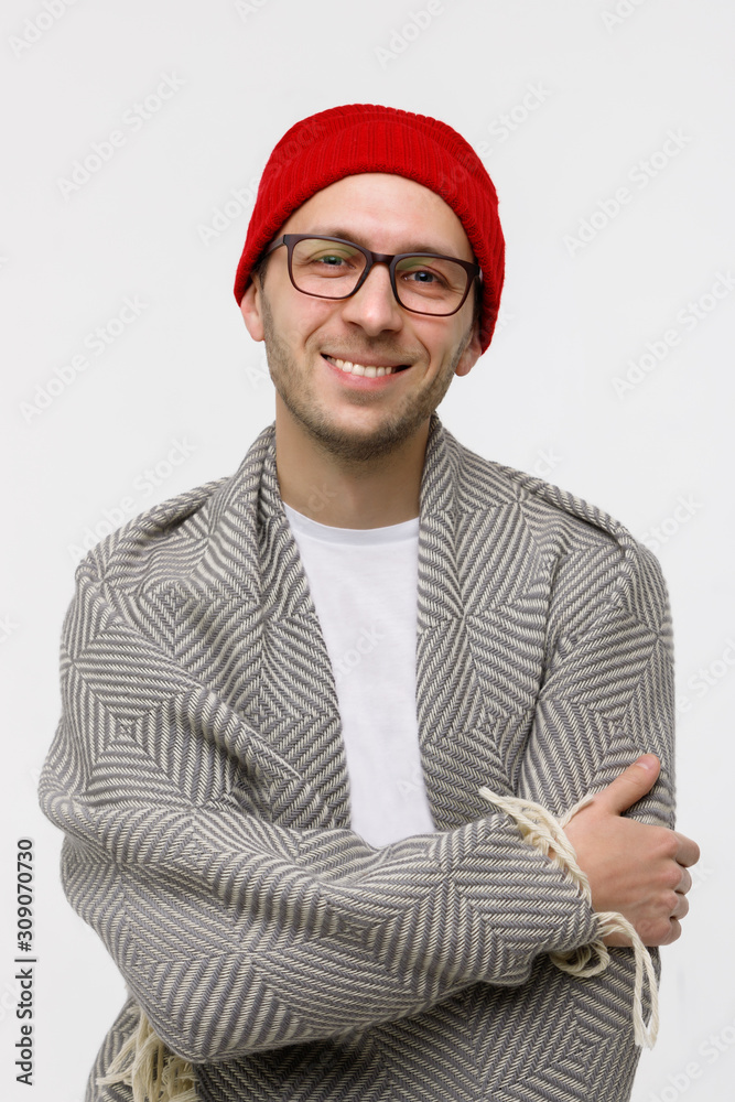 Studio portrait of cheerful stylish hipster guy with light bristles on the face smiling at camera, wearing glasses, red beanie and wrapped in plaid, arms crossed. Isolated on white background. Studio portrait of cheerful stylish hipster guy with light bristles on the face smiling at camera, wearing glasses, red beanie and wrapped in plaid, arms crossed. Isolated on white background.