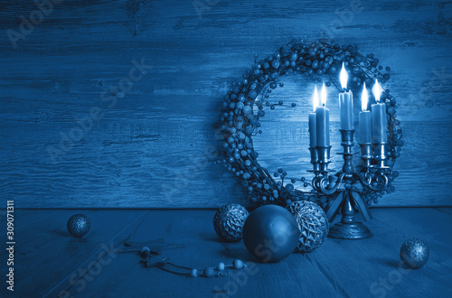 Trendy blue monochrome Xmas composition with candles in candelabrum, berry wreath and classic vintage glass trinkets. Vintage Christmas decorations on wood with copy-space.