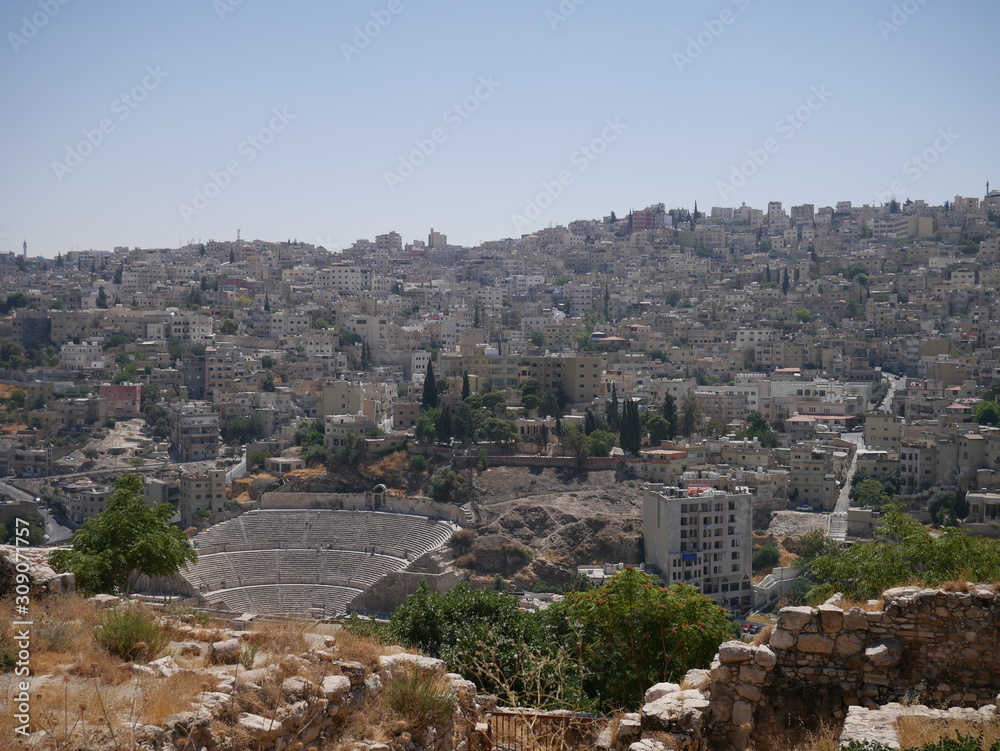 Cityscape of Amman, capitol of Jordan, grey panorama of a modern Arabic city with improvised houses on a hill between few green trees and a Roman theatre under the blue sky