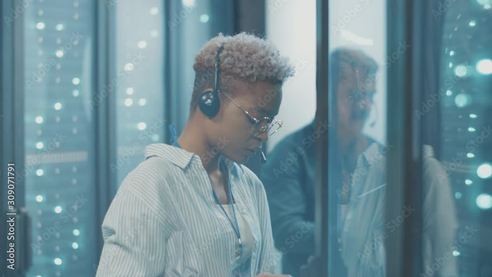 Black IT technician woman receiving online support inspecting an open server cabinet transferring network data into laptop collaborating at cyber security.