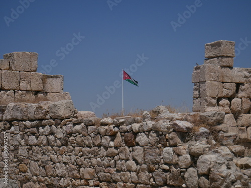 Jordanian flag flying over Roman ruins of the citadel of Amman, capitol of Jordan, remains of a city build from stone and tall pillars on a brown hill in the middle of a city