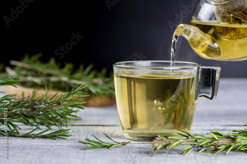 Glass cup of healthy rosemary tea pouring from teapot with fresh rosemary bunch on white wooden rustic background, winter herbal hot drink concept, salvia rosmarinus photo