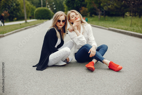 Pretty girls in a summer park. Friends sitting on a asphalt. Ladies with blonde hair. Woman in a sunglasses. © hetmanstock2