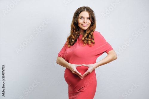 Beautiful pregnant young woman standing over grey wall, making heart shape with her hands. Copy space. Happy pregnancy concept. 