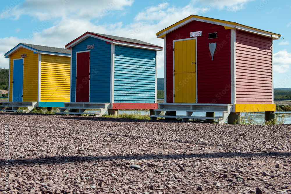 Red, blue and yellow sheds with colorful doors lined up on a beach with blue sky and clouds in the background.