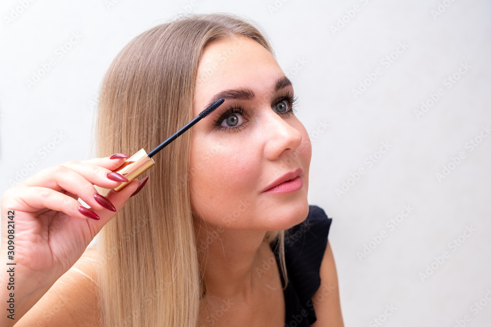 Headshot of female with bright makeup applying mascara on eyelashes.