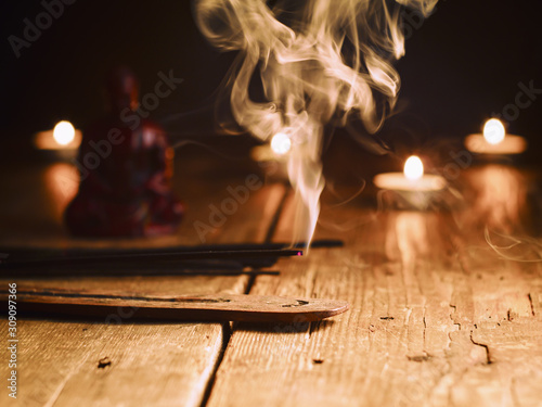 Smoking incense stick in the foreground. On background Small statue of Buddha with incense sticks and burning candles photo