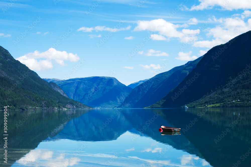 Small wooden boat moored to a red buoy floats on waters of the Lustrafjord in Norway, with reflections of mountains, blue sky and fluffy white clouds. Beautiful Europe Tourism Scene.
