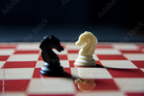 Knight vs knight. White knight in focus vs black knight on chess board. Reflection of white knight on chess board
