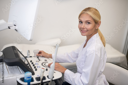 Blond pleasant ultrasound specialist in white robe sitting at the ultrasound machine