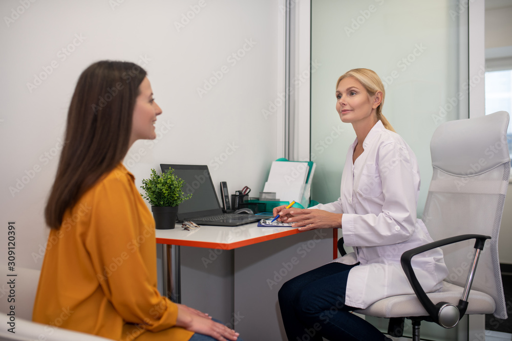 Fototapeta premium Blond doctor in white robe talking to her young female patient