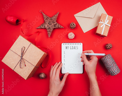Blank notebook, pen, craft gift boxes, envelope, red bow, clew of tricolor rope, ribbon and decorations over red festive background. New year planning.