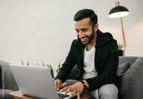 Trendy handsome Gen z Indian arabic man working on laptop in waiting area, Smiling and staring at screen