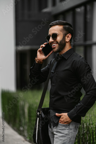 Trendy handsome Gen z Indian arabic man talking on mobile phone, laughing and smiling, looking into the distance