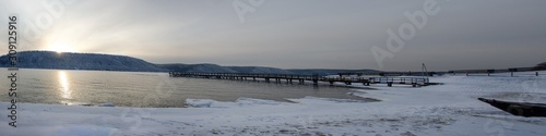 Panorama. Old pier on the river in the winter season. The river has not frozen. Sunny sunset on the river. In the background is a winter forest. Color trend year 2020.
