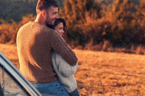 Beautiful young couple enjoying time together outdoor sitting on meadow leaning on old fashioned car embracing each other.