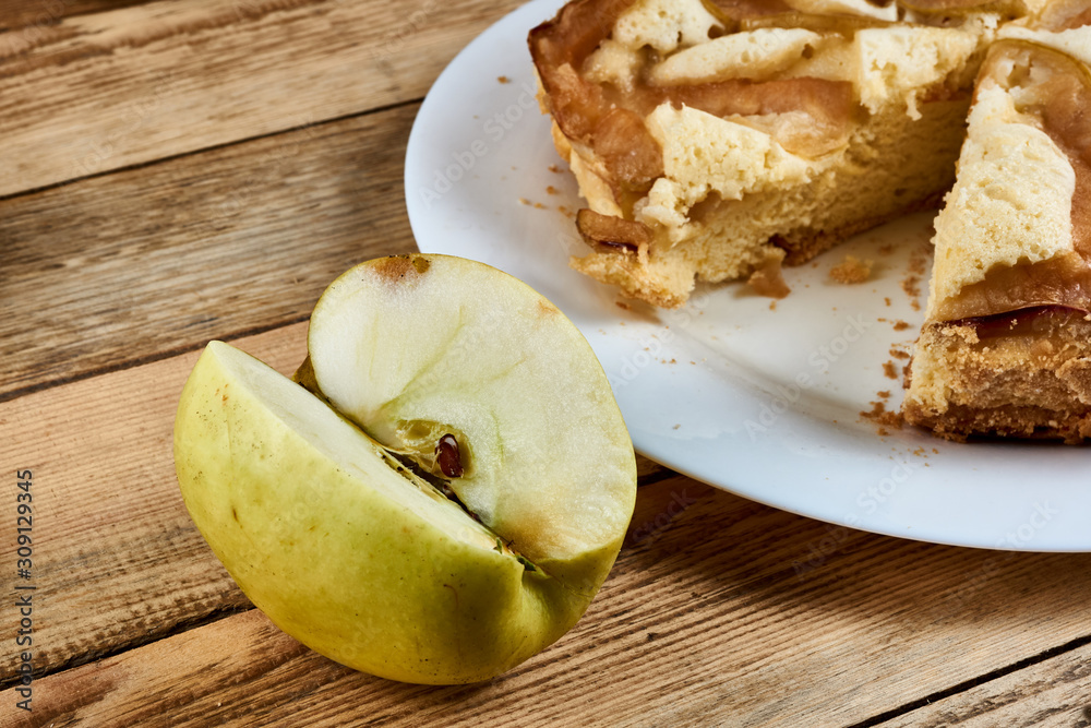 Sliced piece of green apple next to a baked apple pie on a white plate on a wooden background