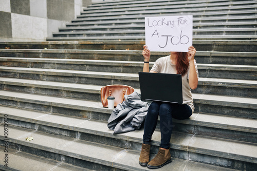 Young woman sitting on steps with laptop and showing board with looking for a job inscription photo