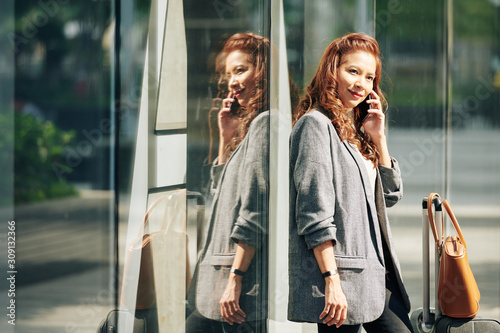 Portrait of smiling pretty Vietnamese woman leaning on shop window when waiting for taxi and calling on phone