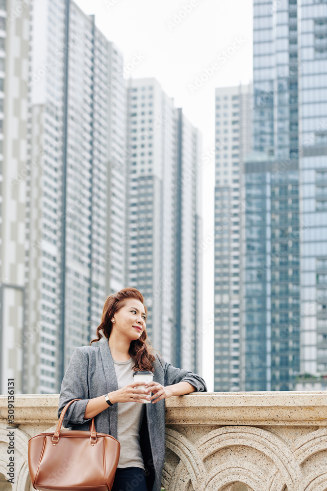 Pretty young Asian businesswoman drinking morning take away coffee when standing on bridge with new district in background