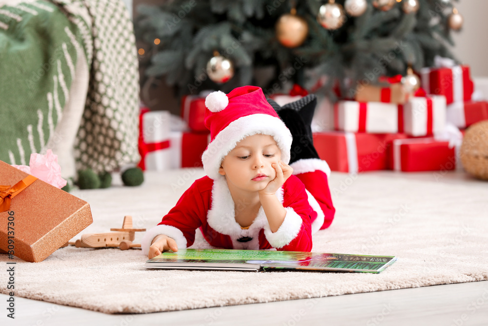 Cute little boy in Santa costume and with book at home on Christmas eve Cute little boy in Santa costume and with book at home on Christmas eve