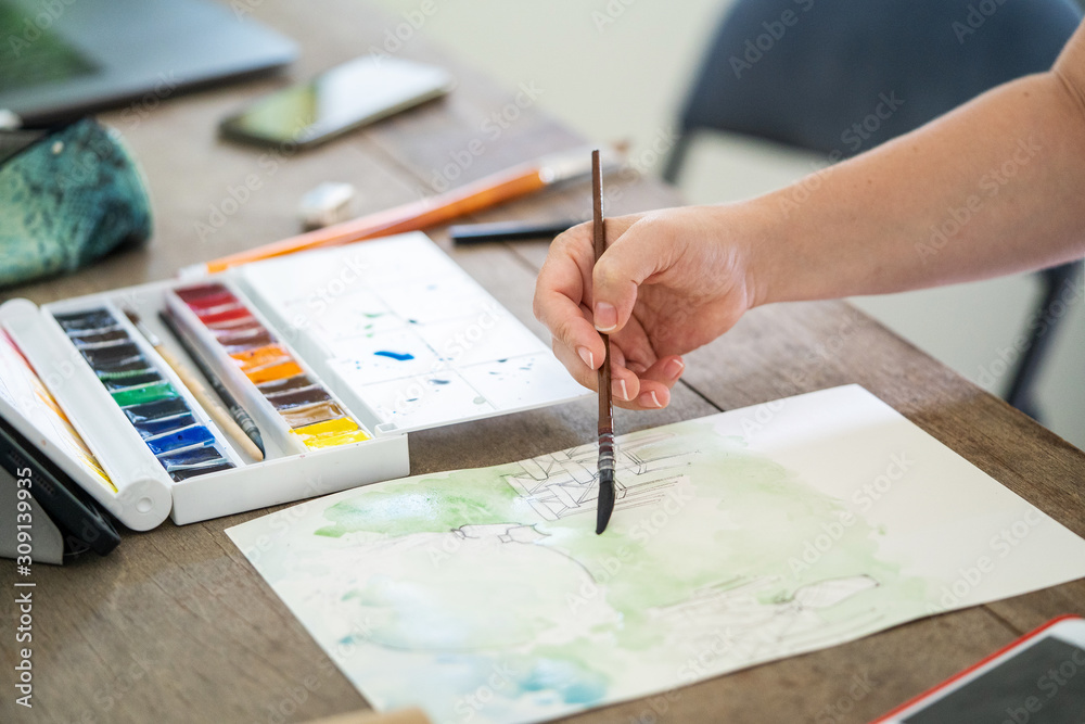 Female hand of an artist with a brush, makes a watercolor drawing on white paper on a wooden table. Female hand of an artist with a brush, makes a watercolor drawing on white paper on a wooden table.