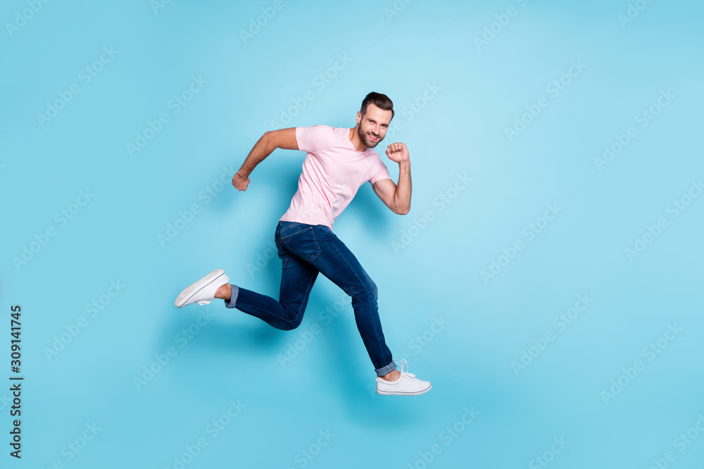 Full length profile photo of attractive guy jumping high sportive competitions participant wear casual pink t-shirt jeans sneakers isolated blue color background Full length profile photo of attractive guy jumping high sportive competitions participant wear casual pink t-shirt jeans sneakers isolated blue color background