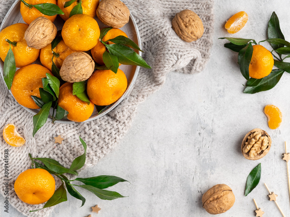 Fresh bright tangerines (oranges) with green leaves and walnuts on gray plate on pullover background, cozy winter mood, top view, frame, template layout